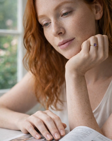 Kobieta o rudych włosach czytająca książkę przy oknie, na dłoni pierścionek z ametystem w złoconej oprawie ByLucy.