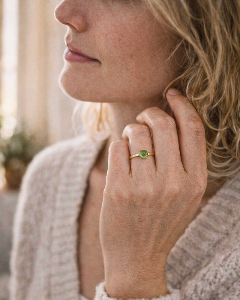 Kobieta w jasnym swetrze dotykająca szyi, nosząca subtelny pierścionek z perydotem w złoconej oprawie, biżuteria ByLucy w naturalnym świetle.