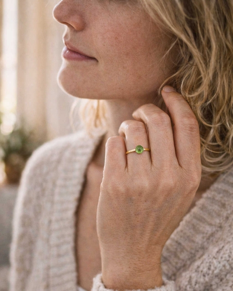 Kobieta w jasnym swetrze dotykająca szyi, nosząca subtelny pierścionek z perydotem w złoconej oprawie, biżuteria ByLucy w naturalnym świetle.